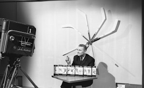 A man is seated at a table with several hockey team display cards in front of him. Behind the man, several hockey sticks are arranged in a fan pattern against a wall. To the left, an old-fashioned television camera is pointed towards the man.