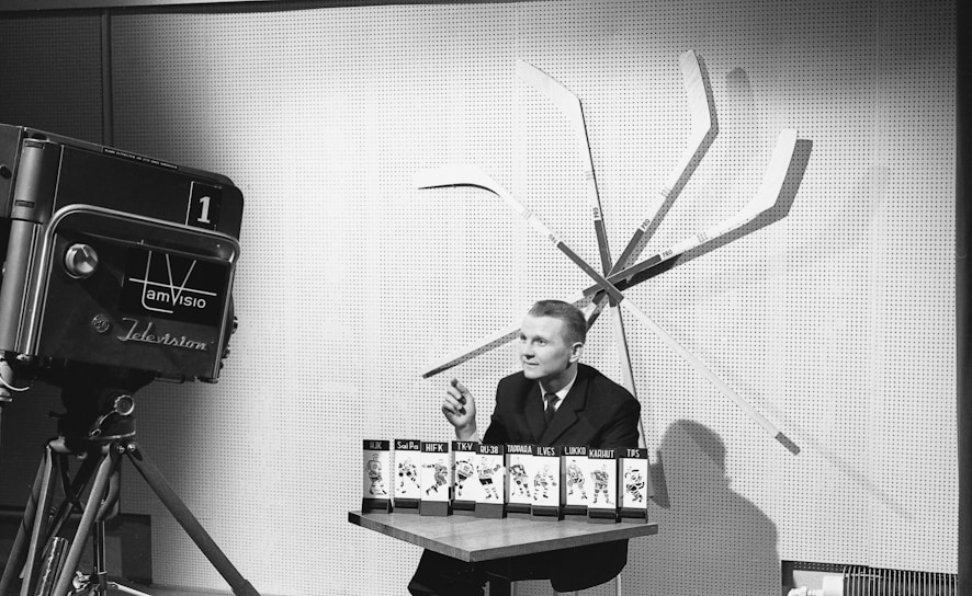 A man is seated at a table with several hockey team display cards in front of him. Behind the man, several hockey sticks are arranged in a fan pattern against a wall. To the left, an old-fashioned television camera is pointed towards the man.
