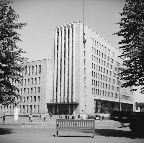 A large, multi-story brick building with a modernist architectural style. The facade features vertical window patterns, and the entrance at ground level is marked by the word 'POST' in large letters. People are walking around the square in front of the building, and trees frame the view on either side.