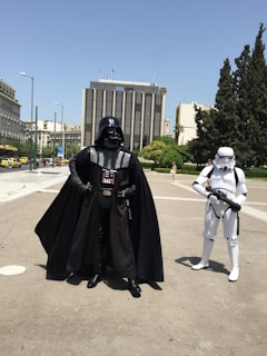 In an urban setting, two people are dressed in iconic costumes representing characters from a popular science fiction franchise, featuring a person in a black armored suit with a cape and helmet, alongside another in a white armored suit with a helmet. They are standing on a paved area in front of a modern building, with some trees and other buildings visible in the background.