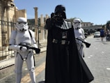 Three individuals dressed in costumes from a popular science fiction franchise are standing in an outdoor urban setting with stone columns and pedestrian walkways. The central figure is in a black outfit with a helmet and cape, flanked by two individuals in white armor and helmets, each holding black blasters.