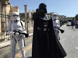 Three individuals dressed in costumes from a popular science fiction franchise are standing in an outdoor urban setting with stone columns and pedestrian walkways. The central figure is in a black outfit with a helmet and cape, flanked by two individuals in white armor and helmets, each holding black blasters.