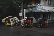 Several motorized tricycles are parked along a wet street in an urban setting. The tricycles have colorful advertisements and decorations on them. A building with a striped awning and various signs is visible in the background, indicating a mix of businesses.