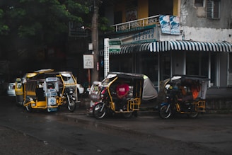 Several motorized tricycles are parked along a wet street in an urban setting. The tricycles have colorful advertisements and decorations on them. A building with a striped awning and various signs is visible in the background, indicating a mix of businesses.