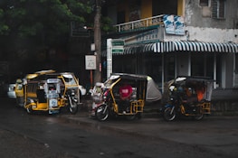 Several motorized tricycles are parked along a wet street in an urban setting. The tricycles have colorful advertisements and decorations on them. A building with a striped awning and various signs is visible in the background, indicating a mix of businesses.