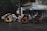 Several motorized tricycles are parked along a wet street in an urban setting. The tricycles have colorful advertisements and decorations on them. A building with a striped awning and various signs is visible in the background, indicating a mix of businesses.