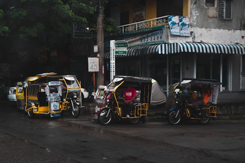 Several motorized tricycles are parked along a wet street in an urban setting. The tricycles have colorful advertisements and decorations on them. A building with a striped awning and various signs is visible in the background, indicating a mix of businesses.