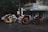 Several motorized tricycles are parked along a wet street in an urban setting. The tricycles have colorful advertisements and decorations on them. A building with a striped awning and various signs is visible in the background, indicating a mix of businesses.
