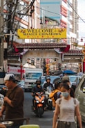 A bustling city street features an archway with a 'Welcome to Manila Chinatown' sign. The area is crowded with cars, motorcycles, and pedestrians. People are engaged in various activities, some wearing masks. The street is lined with buildings, and there are numerous overhead wires.