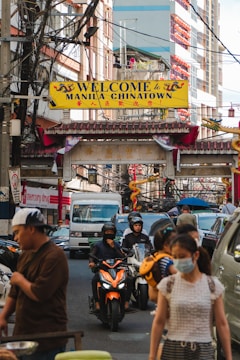 A bustling city street features an archway with a 'Welcome to Manila Chinatown' sign. The area is crowded with cars, motorcycles, and pedestrians. People are engaged in various activities, some wearing masks. The street is lined with buildings, and there are numerous overhead wires.
