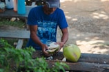 A person is cutting open a green coconut on a wooden bench, surrounded by greenery. The individual is wearing a blue shirt and a cap. A whole coconut and some coconut pieces are visible on the table.