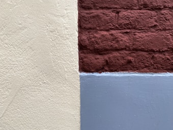 A textured wall with sections painted in three different colors: cream, dark red, and gray. The cream section has a rough surface, the dark red area is made of bricks, and the gray section appears smooth.