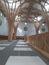 The image depicts the interior of a modern architectural structure featuring intricate wooden beams that form a geometric pattern on the ceiling. The room has a row of rectangular mats on the floor, typical of a prayer space. The walls are adorned with circular calligraphy art. Natural light filters through circular skylights, creating a serene atmosphere.