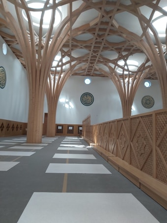 The image depicts the interior of a modern architectural structure featuring intricate wooden beams that form a geometric pattern on the ceiling. The room has a row of rectangular mats on the floor, typical of a prayer space. The walls are adorned with circular calligraphy art. Natural light filters through circular skylights, creating a serene atmosphere.