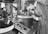 A person is intently examining a precision device or piece of equipment in a laboratory or industrial setting. The individual is wearing a formal outfit, which suggests a professional or scientific context. Various technical instruments surround the workspace, including control panels and gauges, indicating an environment focused on detailed and delicate work.
