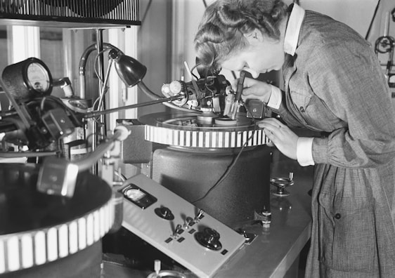 A person is intently examining a precision device or piece of equipment in a laboratory or industrial setting. The individual is wearing a formal outfit, which suggests a professional or scientific context. Various technical instruments surround the workspace, including control panels and gauges, indicating an environment focused on detailed and delicate work.