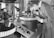A person is intently examining a precision device or piece of equipment in a laboratory or industrial setting. The individual is wearing a formal outfit, which suggests a professional or scientific context. Various technical instruments surround the workspace, including control panels and gauges, indicating an environment focused on detailed and delicate work.