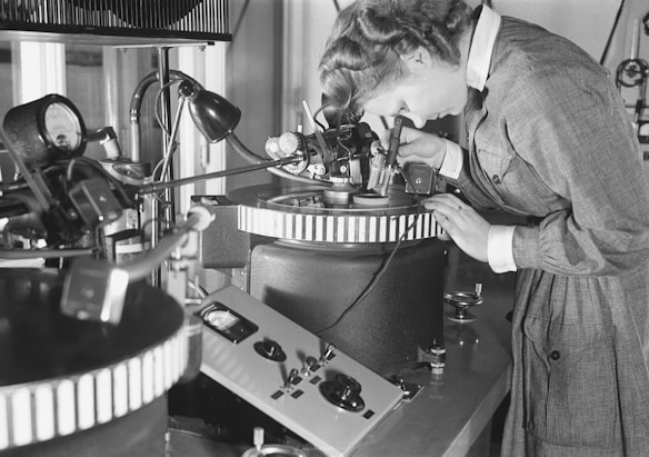 A person is intently examining a precision device or piece of equipment in a laboratory or industrial setting. The individual is wearing a formal outfit, which suggests a professional or scientific context. Various technical instruments surround the workspace, including control panels and gauges, indicating an environment focused on detailed and delicate work.