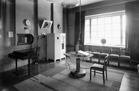 A vintage recording studio features a central microphone suspended from the ceiling, a classic piano to the left, and a small table with a chair. The studio has a large window with curtains, letting in light, and several chairs arranged in a row along the wall. A clock is mounted on the wall above a small window and a cabinet, contributing to the historical atmosphere.