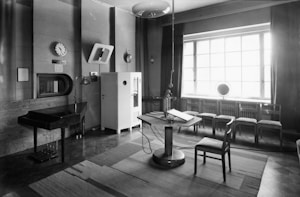 A vintage recording studio features a central microphone suspended from the ceiling, a classic piano to the left, and a small table with a chair. The studio has a large window with curtains, letting in light, and several chairs arranged in a row along the wall. A clock is mounted on the wall above a small window and a cabinet, contributing to the historical atmosphere.