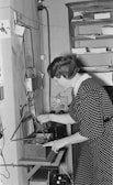 A woman in a polka dot dress is operating an old-fashioned recording or communication device mounted on a wall. She is adjusting a component in a small wooden box with a circular speaker and a triangular stand. Papers and electronic components are visible around the setup, suggesting a vintage office or laboratory setting.