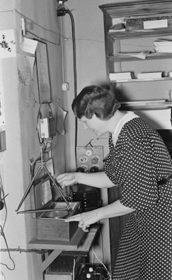 A woman in a polka dot dress is operating an old-fashioned recording or communication device mounted on a wall. She is adjusting a component in a small wooden box with a circular speaker and a triangular stand. Papers and electronic components are visible around the setup, suggesting a vintage office or laboratory setting.