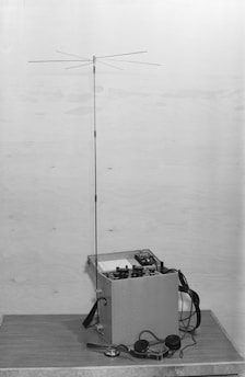A vintage communication device with an extended antenna mounted on a square base. The device features several knobs and dials on its surface, likely for tuning and operation. Attached are headphones and cables, resting on a wooden surface. The background is plain and light-colored.