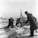 Workers are engaged in manual labor, stripping logs on an icy dock with a large crane structure in the background. The environment appears industrial with a body of water and buildings visible in the distance. Two men with tools focus on removing the bark from large logs, while another man in a dark coat and hat observes or directs the activity. The scene captures a moment of hard physical work in a wintry setting.