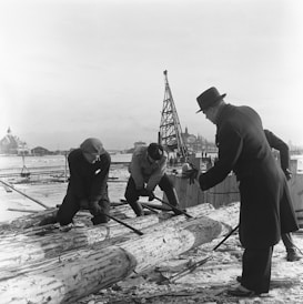 Workers are engaged in manual labor, stripping logs on an icy dock with a large crane structure in the background. The environment appears industrial with a body of water and buildings visible in the distance. Two men with tools focus on removing the bark from large logs, while another man in a dark coat and hat observes or directs the activity. The scene captures a moment of hard physical work in a wintry setting.