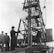 A group of men are gathered around a large, metal drilling rig. One man in a suit and hat appears to be overseeing the operation, holding a microphone or similar device. The rig is actively releasing steam or smoke, indicating ongoing activity. The environment suggests an industrial or construction setting.