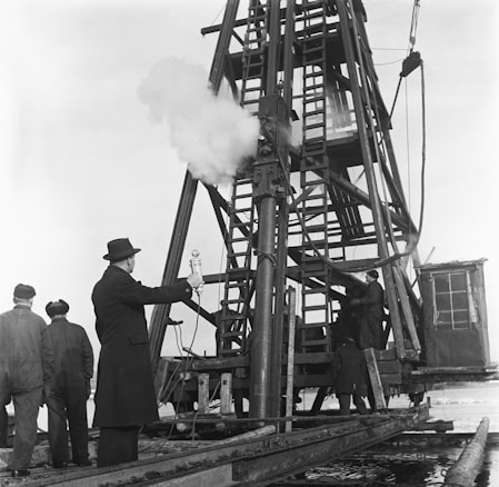 A group of men are gathered around a large, metal drilling rig. One man in a suit and hat appears to be overseeing the operation, holding a microphone or similar device. The rig is actively releasing steam or smoke, indicating ongoing activity. The environment suggests an industrial or construction setting.