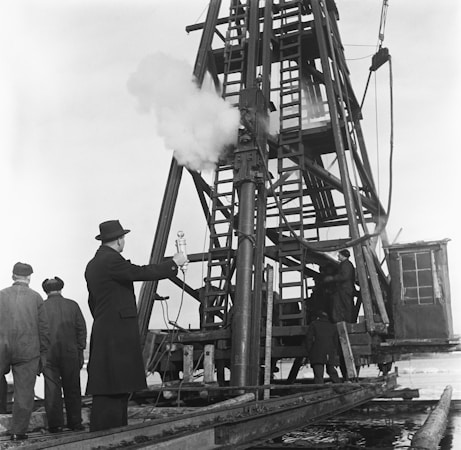 A group of men are gathered around a large, metal drilling rig. One man in a suit and hat appears to be overseeing the operation, holding a microphone or similar device. The rig is actively releasing steam or smoke, indicating ongoing activity. The environment suggests an industrial or construction setting.