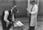 Two men are engaged in some form of scientific experiment or demonstration. One man, dressed in a fedora and vest, is using a saw on a block of material. The other man, wearing a white lab coat, is observing and holding a scientific device similar to a microphone or a measuring instrument. They are standing outside in front of large, patterned doors.