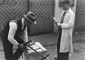 Two men are engaged in some form of scientific experiment or demonstration. One man, dressed in a fedora and vest, is using a saw on a block of material. The other man, wearing a white lab coat, is observing and holding a scientific device similar to a microphone or a measuring instrument. They are standing outside in front of large, patterned doors.