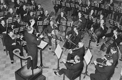 A large orchestra is performing, with a conductor standing on a podium leading the musicians. The orchestra consists of string players, including violinists and cellists, who are seated with music stands in front of them.