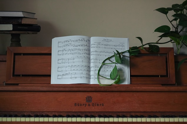 A vintage wooden piano with sheet music open on its stand. A green plant vine elegantly drapes across the music. A stack of books is visible on a shelf in the background.