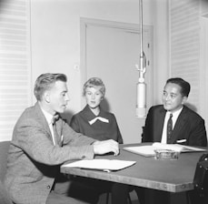 Three individuals are seated around a table with a microphone hanging above them, engaged in a conversation. The setting appears to be a studio or interview room, indicated by the large microphone and professional attire of the participants. A man in a suit and tie, a woman wearing a dress, and another man in a checked suit jacket are present. Books or documents are open in front of them, suggesting a formal discussion or interview.
