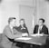 Three individuals are seated around a table with a microphone hanging above them, engaged in a conversation. The setting appears to be a studio or interview room, indicated by the large microphone and professional attire of the participants. A man in a suit and tie, a woman wearing a dress, and another man in a checked suit jacket are present. Books or documents are open in front of them, suggesting a formal discussion or interview.