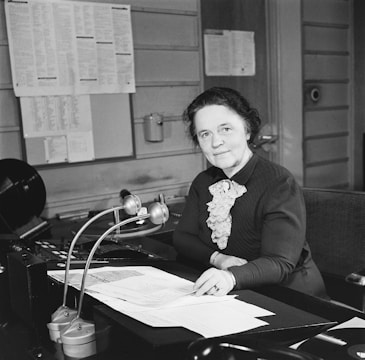 A woman is seated at a desk in an office environment, looking towards the camera. Papers are spread out in front of her, and a phone and other office equipment are visible. The walls are decorated with various papers and notices.