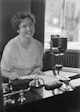 Photo of a smiling woman sitting at a desk with paperwork and a laptop.