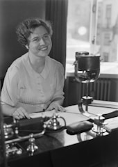 A woman with short curly hair and a button-up shirt is seated at a desk, smiling and looking slightly upward. The desk holds a vintage office machine, papers, and a telephone. A window in the background lets in natural light, revealing a blurred cityscape.