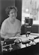 Photo of a smiling woman sitting at a desk with paperwork and a laptop.