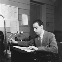 A man wearing a suit sits at a desk, concentrating on the documents in front of him. The desk has a microphone, a telephone, and some papers. The background includes a wall with several sheets of paper posted on it and a door.