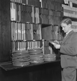 A man dressed in a suit examines film reels in a workspace filled with organized shelves. The shelves are stacked with numerous film cans and file holders.