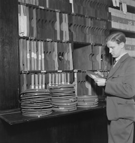A man dressed in a suit examines film reels in a workspace filled with organized shelves. The shelves are stacked with numerous film cans and file holders.