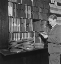 A man dressed in a suit examines film reels in a workspace filled with organized shelves. The shelves are stacked with numerous film cans and file holders.
