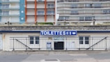 A public restroom facility with a prominent blue and white sign labeled 'TOILETTES' above the entrance. The building features a stone facade and is located at the base of a large apartment complex. The apartments have colorful facades with blue, orange, and beige sections.