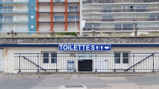 A public restroom facility with a prominent blue and white sign labeled 'TOILETTES' above the entrance. The building features a stone facade and is located at the base of a large apartment complex. The apartments have colorful facades with blue, orange, and beige sections.