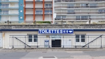 A public restroom facility with a prominent blue and white sign labeled 'TOILETTES' above the entrance. The building features a stone facade and is located at the base of a large apartment complex. The apartments have colorful facades with blue, orange, and beige sections.