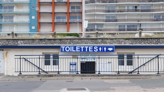 A public restroom facility with a prominent blue and white sign labeled 'TOILETTES' above the entrance. The building features a stone facade and is located at the base of a large apartment complex. The apartments have colorful facades with blue, orange, and beige sections.